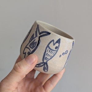 May include: A small, speckled, off-white ceramic cup featuring hand-drawn dark blue fish illustrations. The cup has a rounded, tapered shape with a smooth rim and a textured, speckled surface finish throughout the exterior of the vessel.