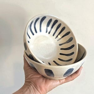 May include: Two stacked ceramic bowls with a cream-coloured glaze and hand-painted dark blue vertical brushstroke patterns. The bowls feature a rounded shape with a smooth finish, showcasing a consistent design on both the interior and exterior surfaces.