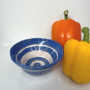 May include: A handcrafted ceramic bowl with a blue exterior and a white interior, showcasing a spiral pattern. An orange and a yellow bell pepper are positioned nearby, forming a vibrant still life composition.