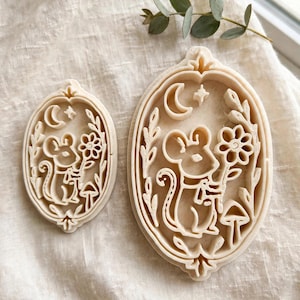 May include: Two off-white oval cookie cutters featuring a raised relief design of a mouse holding a flower, accompanied by a mushroom, a crescent moon, and a small star. The cutters have a decorative border and are made of a solid, light-colored material.