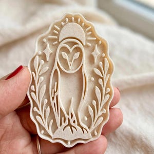 May include: A cream-colored, speckled plastic cookie cutter featuring a detailed embossed design of a barn owl centered within a scalloped frame. The owl is flanked by leafy branches and topped with a crescent moon and small star accents.