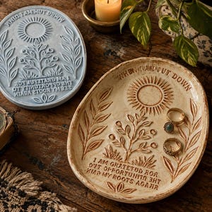 May include: Two oval ceramic trinket dishes featuring embossed designs of a sun, a tree, and foliage. The text reads, "WHEN I AM CUT DOWN, I AM GRATEFUL FOR THE OPPORTUNITY TO SEE MY ROOTS AGAIN." One dish is light blue, and the other is a cream color.