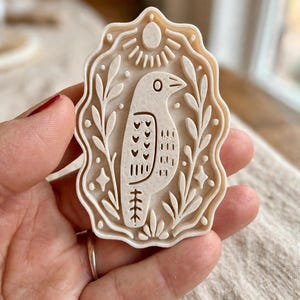 May include: A cream-colored, rectangular cookie cutter with a scalloped edge featuring an embossed design of a bird facing right, surrounded by stylized leafy branches, small dots, and a sun motif at the top, all rendered in a raised relief pattern.