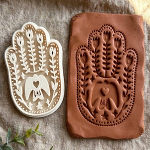 May include: A cream-colored Hamsa hand stamp featuring a floral and bird design with a central heart, positioned next to a rectangular slab of terracotta-colored clay that has been impressed with the same Hamsa hand pattern.