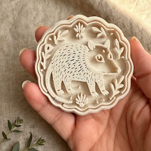 May include: A round, off-white cookie cutter featuring a detailed embossed design of an opossum surrounded by small, stylized leaf and floral motifs. The cutter has a scalloped edge and a textured surface for creating impressions in dough.