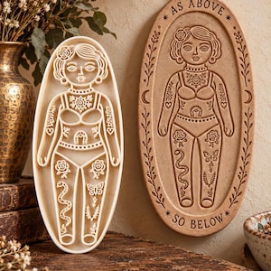 May include: An oval-shaped cream-colored stamp cutter and a matching tan oval plaque featuring an embossed figure with intricate floral, snake, and butterfly designs. The plaque includes the text AS ABOVE and SO BELOW with crescent moon symbols.