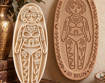 May include: An oval-shaped cream-colored stamp cutter and a matching tan oval plaque featuring an embossed figure with intricate floral, snake, and butterfly designs. The plaque includes the text AS ABOVE and SO BELOW with crescent moon symbols.