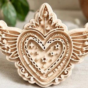 May include: A light-colored wooden cookie cutter featuring a central heart shape with wings extending from both sides. The design includes intricate cutouts of stars, small crescents, and decorative scalloped borders within the heart and wings.