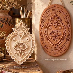 May include: A cream-colored heart-shaped stamp cutter featuring an eye and flame design, alongside a matching oval terracotta wall plaque with the text AS WITHIN SO WITHOUT, both displaying intricate embossed details and decorative floral borders.
