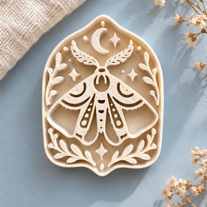May include: A cream-colored, arch-shaped cookie cutter featuring an embossed design of a moth centered beneath a crescent moon and stars, framed by delicate leafy branches on both sides, crafted with a raised edge for cutting.