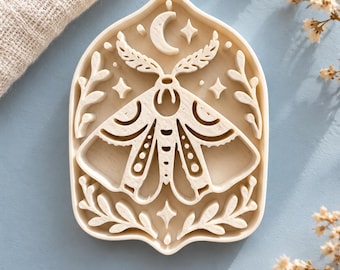 May include: A cream-colored, arch-shaped cookie cutter featuring an embossed design of a moth centered beneath a crescent moon and stars, framed by delicate leafy branches on both sides, crafted with a raised edge for cutting.