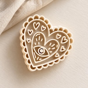 May include: A beige heart-shaped cookie cutter featuring a scalloped edge and an interior design of a central eye symbol surrounded by smaller heart shapes and short radiating lines, crafted from a solid, smooth, light-colored material.
