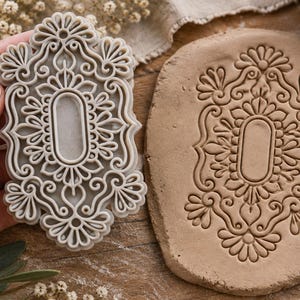 May include: A light gray rectangular clay stamp featuring an intricate, symmetrical filigree pattern with a central oval cutout, shown alongside a piece of tan clay that has been impressed with the same detailed decorative design.