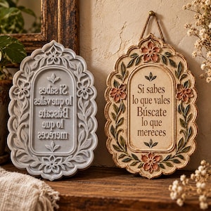 May include: Two oval wall plaques with ornate floral borders. The right plaque is cream-colored with pink flowers and green leaves, featuring the text "Si sabes lo que vales Búscate lo que mereces". The left plaque is a grey mirror-image version.
