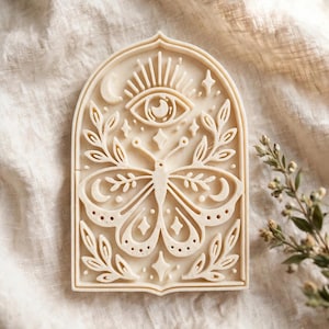 May include: A cream-coloured arch-shaped cookie cutter featuring an embossed design of a central butterfly surrounded by leafy branches, a crescent moon, a sunburst, and an eye symbol, all contained within a decorative arched frame.
