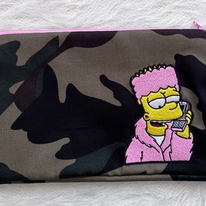 May include: A camouflage fabric zippered pouch with a pink embroidered patch of a cartoon character with pink hair, a yellow face, and a pink jacket talking on a phone.