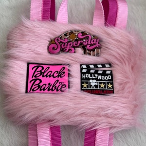 May include: Pink faux fur backpack with black and pink patches that say "Black Barbie" and "Hollywood Lights Camera Action". The backpack also has a patch that says "Superstar" in pink letters.
