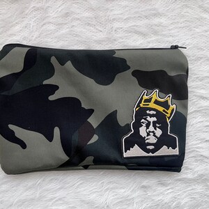 May include: Camouflage print zippered pouch featuring a black and gold embroidered patch of a crowned figure. The pouch has a black zipper and a pattern of black, gray, and olive green shapes.
