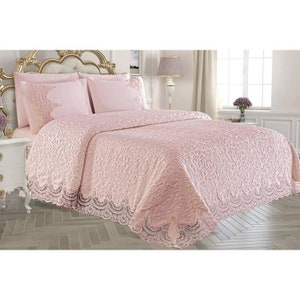 May include: A light pink bedspread and two matching pillow shams featuring an intricate embossed swirl pattern across the surface. The edges of the bedspread and the pillow shams are finished with a wide, decorative lace trim in the same color.