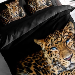 May include: A black bedding set featuring a large, high-definition photographic print of a leopard with striking blue eyes. The design includes two matching pillowcases and a duvet cover, all displaying the same leopard portrait against a black background.