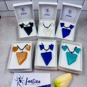 May include: Six sets of jewellery, each containing a triangular pendant and matching earrings on a black cord, presented in white boxes. The sets feature various colours including black, white, blue, orange, and teal, with a logo card labelled Jantina.