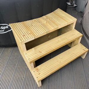May include: Wooden steps with three tiers leading up to a hot tub. The steps are made of light brown wood and have a slightly curved shape. The steps are sitting on a black, textured surface.