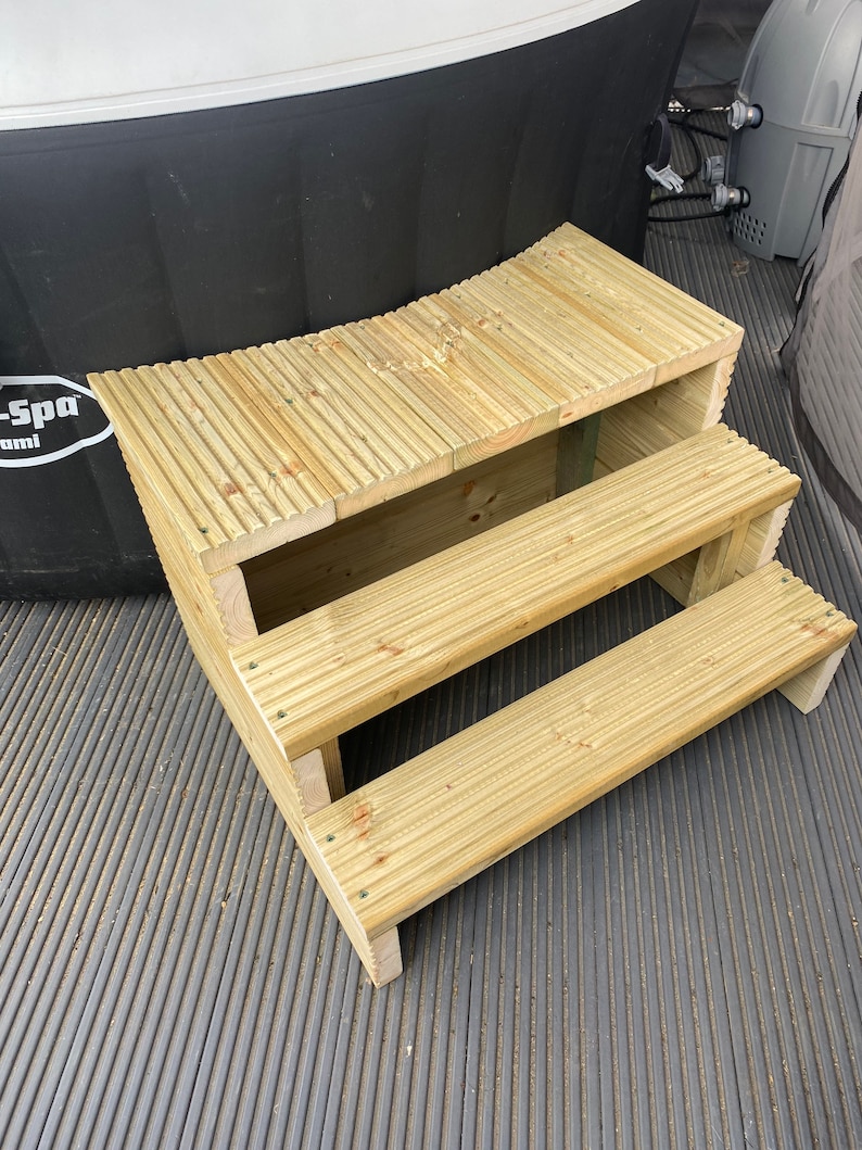 May include: Wooden steps with three tiers leading up to a hot tub. The steps are made of light brown wood and have a slightly curved shape. The steps are sitting on a black, textured surface.