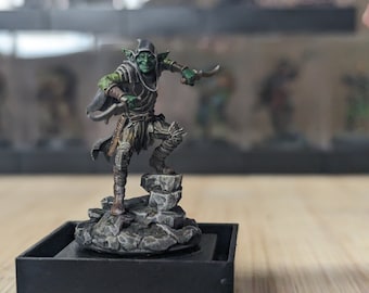 May include: A green goblin miniature figure with a black hooded cloak, holding a dagger, standing on a rocky base.