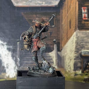 May include: A detailed miniature figurine of a warrior in a dynamic pose, holding a spear. The figure is clad in armor with red and black accents, and a helmet. The base is a dark square, and the background depicts a rustic village scene.