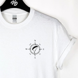 May include: White t-shirt with a black compass design featuring a paraglider inside. The compass points are labelled N, S, E, and W.