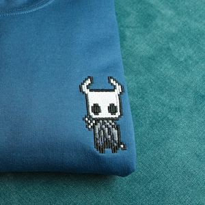 May include: A folded, teal-blue sweatshirt featuring a pixelated character design on the front. The design is a white and black character with a horned head. The sweatshirt is made of a soft material.