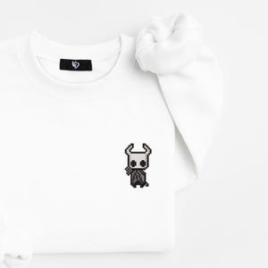 May include: White sweatshirt with a small, pixelated black and white character design on the front. The sweatshirt has long sleeves and a crew neck. A small black tag is visible near the neckline.