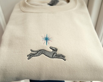Pode incluir: Uma camisa de moletom de cor creme com um cão bordado em cinza correndo com uma estrela azul e rosa acima.