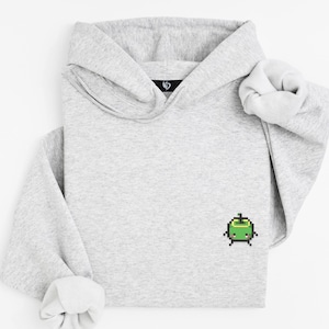 Könnte beinhalten: Ein hellgrauer Kapuzenpullover mit einem pixeligen grünen Charakter auf der Vorderseite. Der Hoodie hat eine Kapuze und lange Ärmel mit gerippten Bündchen. Das Kleidungsstück ist aus einem weichen, bequemen Material gefertigt.