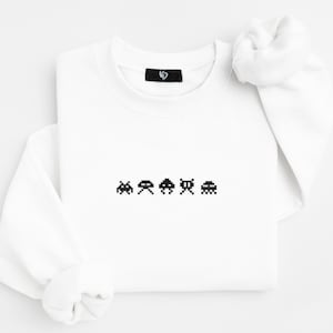 May include: White crew neck sweatshirt with a pixelated video game graphic across the chest. The graphic features four different alien-like characters in black. The sweatshirt has long sleeves and a small black tag near the neckline.