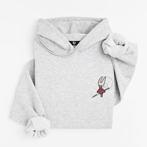 May include: A light gray hoodie with a small embroidered design of a character holding a needle. The hoodie has a hood and long sleeves with folded cuffs. The design is in red, black, and white.