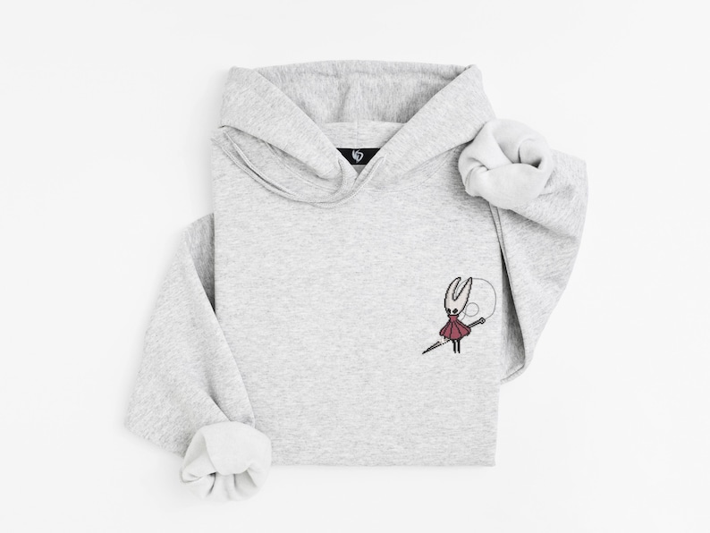 May include: A light gray hoodie with a small embroidered design of a character holding a needle. The hoodie has a hood and long sleeves with folded cuffs. The design is in red, black, and white.