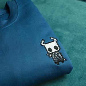 May include: Teal blue sweatshirt with a pixelated embroidered design of a character from the video game Hollow Knight. The design is in black, white, and gray. The sweatshirt is folded on a teal green surface.