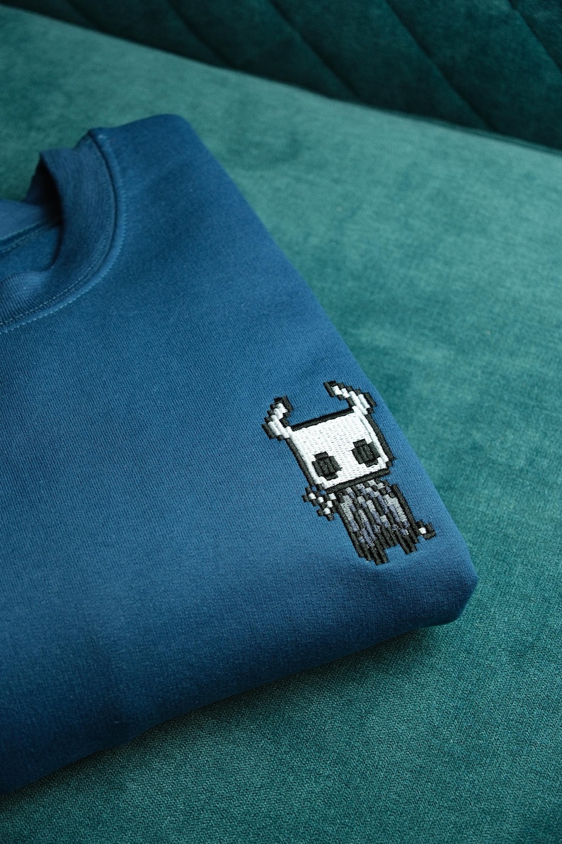 May include: Teal blue sweatshirt with a pixelated embroidered design of a character from the video game Hollow Knight. The design is in black, white, and gray. The sweatshirt is folded on a teal green surface.