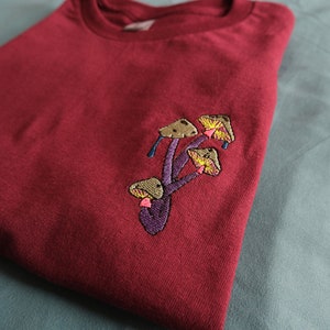 Peut inclure: Un t-shirt marron avec un motif brod&eacute; de champignons en nuances de brun, jaune, rose et violet.