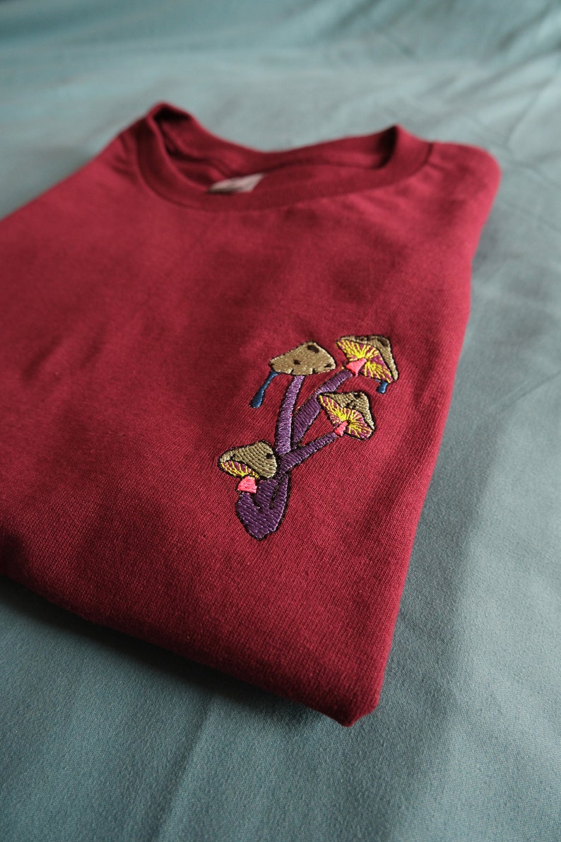 Peut inclure: Un t-shirt marron avec un motif brod&eacute; de champignons en nuances de brun, jaune, rose et violet.