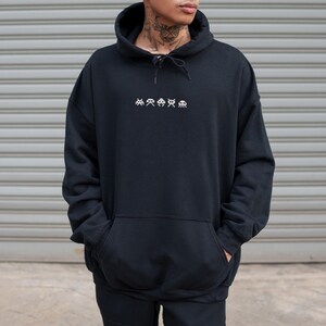 May include: Black hoodie with a drawstring hood and a kangaroo pocket. The front of the hoodie features a white embroidered design of pixelated alien figures. The person is wearing the hoodie and has their hands in the pockets.
