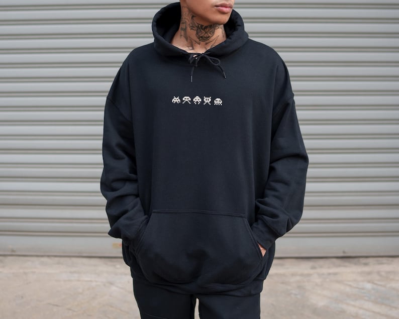 May include: Black hoodie with a drawstring hood and a kangaroo pocket. The front of the hoodie features a white embroidered design of pixelated alien figures. The person is wearing the hoodie and has their hands in the pockets.
