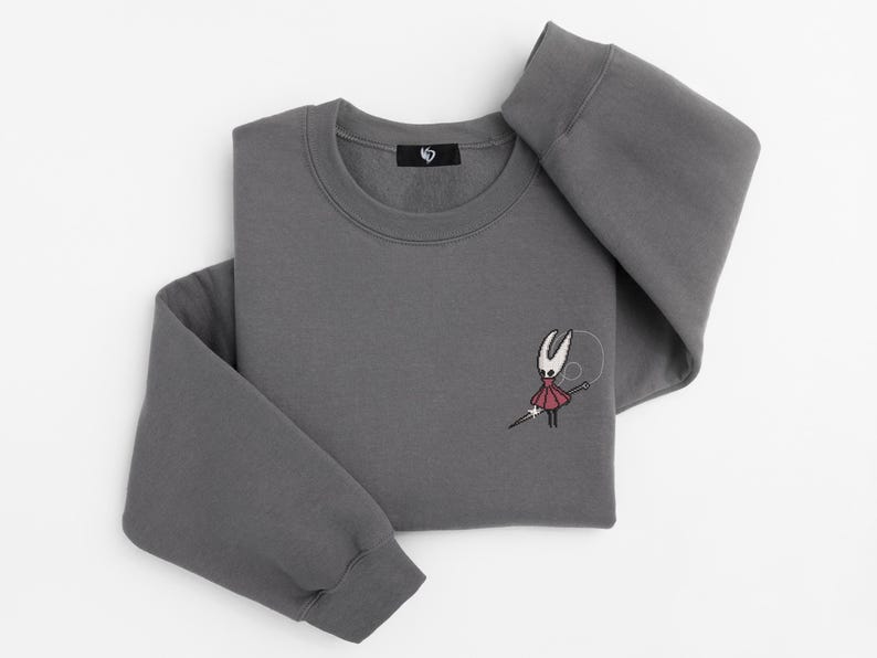 May include: A charcoal gray sweatshirt with a crew neck and long sleeves. The sweatshirt features an embroidered design of a character holding a needle. A small black tag is sewn on the neckline.