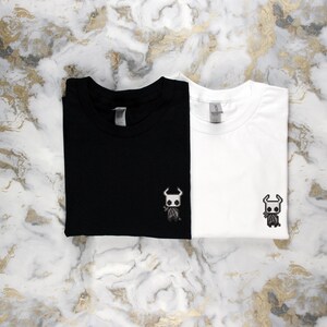 May include: Two folded t-shirts, one black and one white, each with a small embroidered character design on the front. The design features a horned figure. The shirts are displayed on a marble-patterned surface.