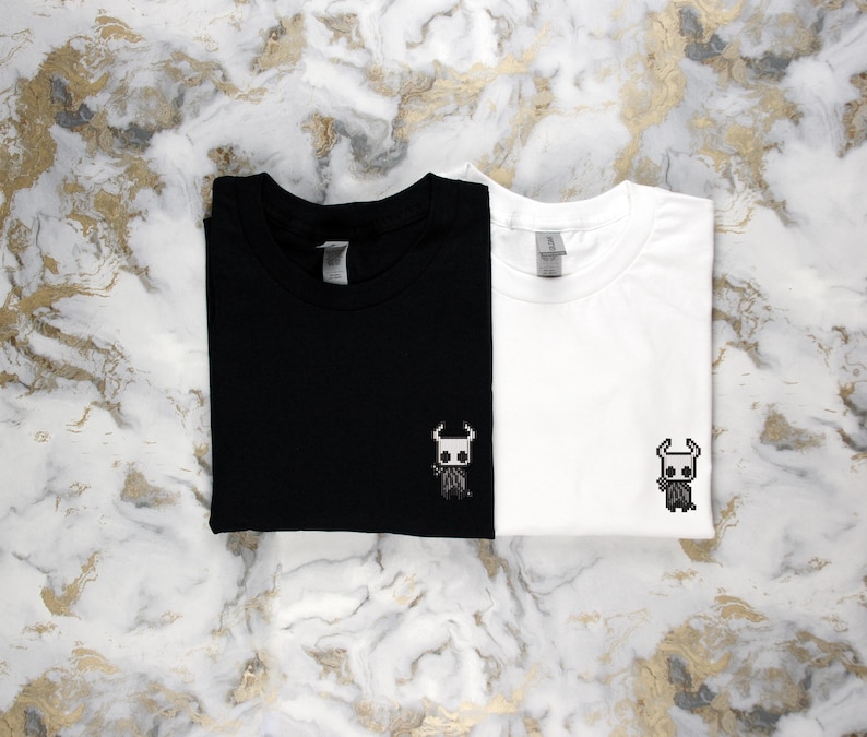 May include: Two folded t-shirts, one black and one white, each with a small embroidered character design on the front. The design features a horned figure. The shirts are displayed on a marble-patterned surface.