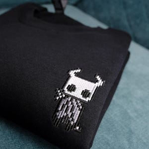 May include: Black sweatshirt featuring an embroidered pixel art design of a character. The design is in shades of white and gray, and is located on the upper left chest area of the sweatshirt. The sweatshirt is folded and resting on a teal surface.