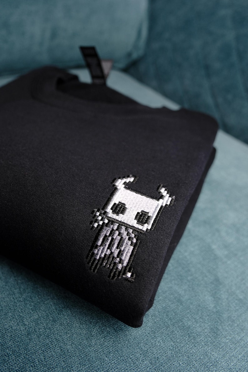 May include: Black sweatshirt featuring an embroidered pixel art design of a character. The design is in shades of white and gray, and is located on the upper left chest area of the sweatshirt. The sweatshirt is folded and resting on a teal surface.