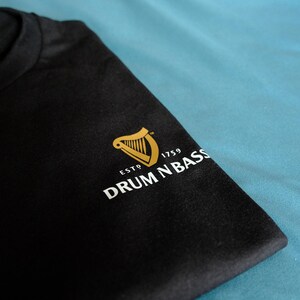 May include: Black t-shirt with a gold harp logo and the text "ESTD 1759 DRUM N BASS" in white.