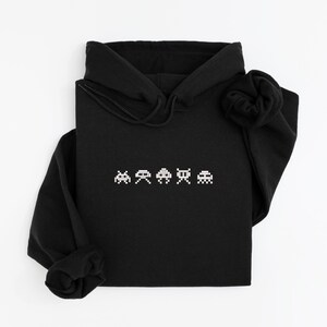 May include: Black hooded sweatshirt with a white pixelated video game graphic across the chest. The graphic features four retro space invader-style characters. The sweatshirt has a drawstring hood and long sleeves.